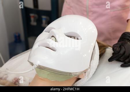 Frau liegt im Bett mit LED-Lichttherapie Gesichtsmaske und entspannen. LED-Lichtmaske für die Gesichtshautbehandlung, franfessionelles Kosmetologieverfahren. Stockfoto