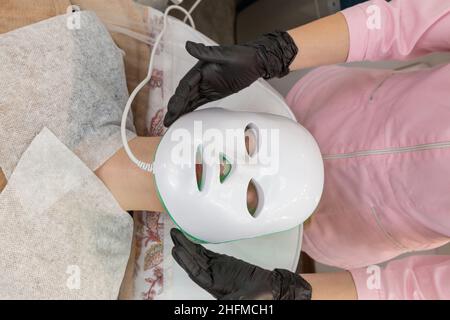 Frau liegt im Bett mit LED-Lichttherapie Gesichtsmaske und entspannen. LED-Lichtmaske für die Gesichtshautbehandlung, franfessionelles Kosmetologieverfahren. Stockfoto