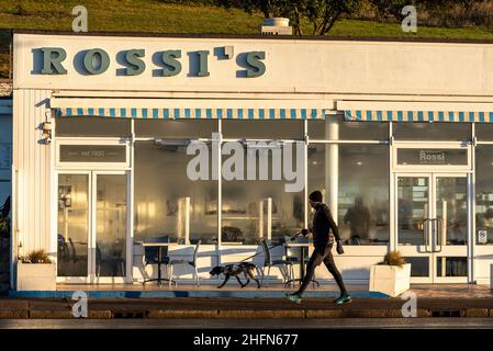 Frühmorgendlicher Hundespaziergänger, der an Rossis Eisdiele auf der Western Esplanade, Southend on Sea, Essex, Großbritannien, vorbeikommt. Sonnenaufgang im Winter. Kondensation an Fenstern Stockfoto