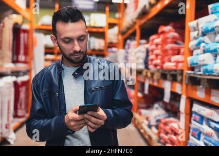 Ein Mann tippt auf einem Smartphone zwischen Reihen von Baumaterialien in einem Baumarkt Stockfoto