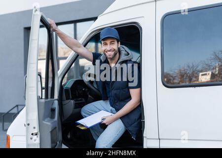 Glücklicher Lieferer, der die Kamera ansah, während er die Zwischenablage und das Paket im Auto hielt Stockfoto