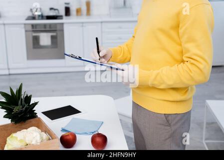 Beschnittene Ansicht des Mannes in Latexhandschuhen, der auf der Zwischenablage neben frischen Lebensmitteln und einem digitalen Tablet in der Küche schreibt Stockfoto