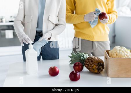 Beschnittene Ansicht eines Paares in Latexhandschuhen, die frische Lebensmittel mit Antiseptikum in der Nähe der Kiste in der Küche reinigen Stockfoto