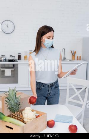 Frau in medizinischer Maske mit digitalen Tablette und nehmen Apfel aus Holzkiste in der Küche Stockfoto