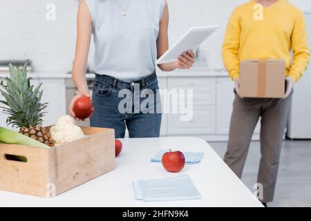 Beschnittene Ansicht einer Frau mit einem digitalen Tablet in der Nähe von frischen Lebensmitteln und eines Freundes mit Karton zu Hause Stockfoto