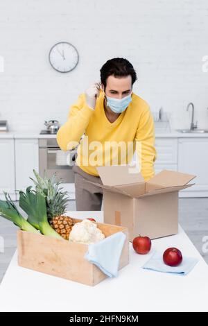 Mann mit medizinischer Maske in der Nähe von frischen Lebensmitteln und Boxen in der Küche Stockfoto