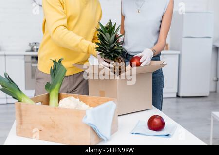 Beschnittene Ansicht eines Paares in Latexhandschuhen, das frisches Obst in der Nähe von Schachteln und Lumpen in der Küche hält Stockfoto