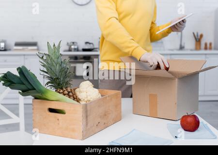 Beschnittene Ansicht des Mannes in Latexhandschuhen mit digitalem Tablet und dem Abnehmen von Apfel aus der Kartonschachtel in der Küche Stockfoto