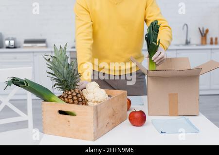 Beschnittene Ansicht eines Mannes in Latexhandschuhen, der frische Lebensmittel in der Nähe von Schachteln und Lumpen in der Küche mitnimmt Stockfoto