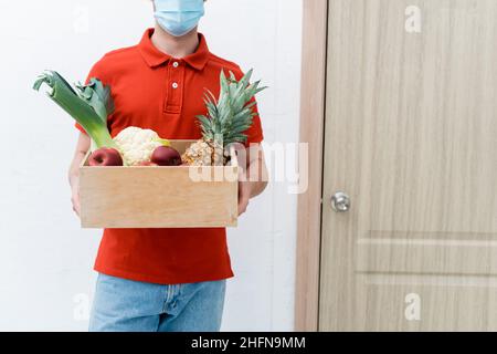 Beschnittene Ansicht des Lieferers in einer Schutzmaske, der eine Holzkiste mit Essen in der Nähe der Tür im Flur hält Stockfoto