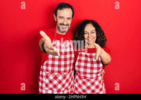 Mittelalterpaar hispanische Frau und Mann tragen Kochschürze lächelnd freundlich mit Handschlag als Begrüßung und Begrüßung. Erfolgreiches Geschäft. Stockfoto