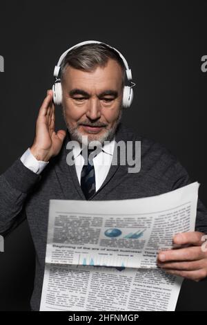 Bärtiger älterer Mann in Kopfhörern, der Zeitung auf dunkelgrau isoliert liest Stockfoto