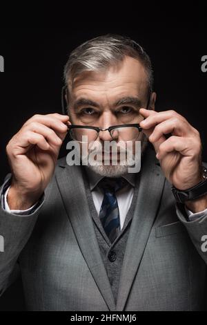 Älterer bärtiger Geschäftsmann, der eine Brille in der Hand hält, während er die Kamera isoliert auf Schwarz anschaut Stockfoto