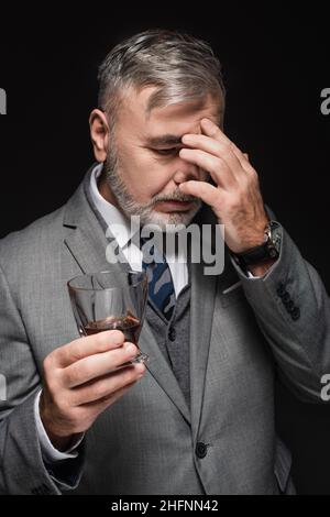 Erschöpfter älterer Mann, der ein Glas Whiskey in der Hand hält, während er an schwarzen Kopfschmerzen leidet Stockfoto