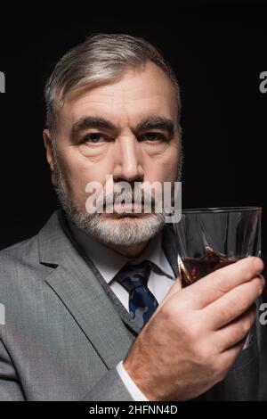 Älterer bärtiger Mann mit Blazer und Krawatte, der ein Glas Whiskey auf Schwarz gehalten hat Stockfoto