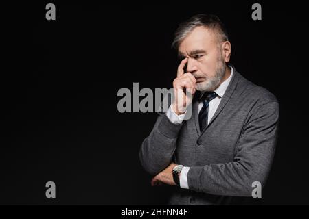 Nachdenklicher älterer Mann in grauem Jumper, der das Gesicht berührt, während er isoliert auf Schwarz denkt Stockfoto