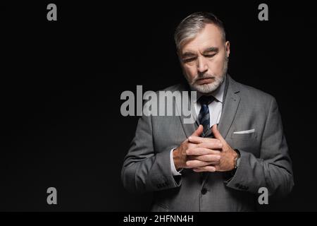 Nachdenklicher älterer Mann in Blazer, der mit zusammengekrallten Händen auf Schwarz isoliert steht Stockfoto