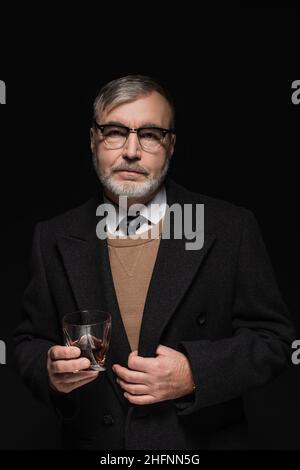 Älterer bärtiger Mann in Mantel und Brille, der ein Glas Whiskey auf Schwarz gehalten hat Stockfoto