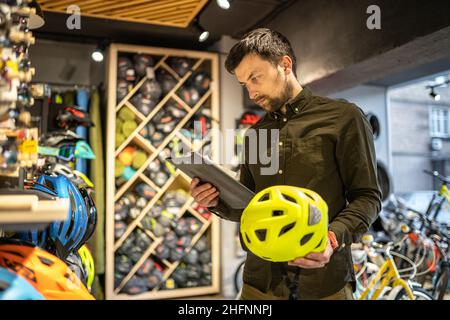 Ein männlicher Bike-Shop-Manager führt in einem Bike-Shop eine Inventarisierung von Sporthelmen durch. Der Besitzer eines Sportladens mit einem Clipboard in den Händen prüft die Stockfoto