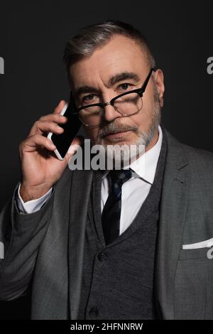 Älterer Mann in Anzug und Brille, der auf die Kamera schaut, während er auf dem Handy spricht, isoliert auf dunkelgrau Stockfoto