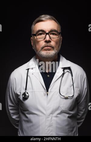 Leitender bärtiger Arzt mit Stethoskop, der die Kamera isoliert auf Schwarz ansieht Stockfoto