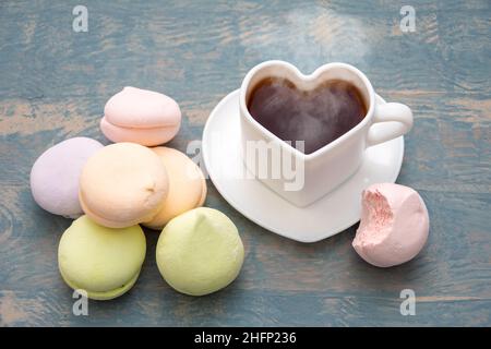 Runde mehrfarbige Marschmeichs in einem Stapel, eine abgebissene Hälfte eines Marschmeiches und eine weiße herzförmige Tasse mit schwarzem dampfendem heißen Kaffee auf einem weißen Stockfoto