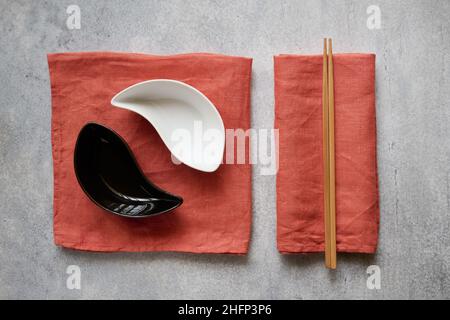 Draufsicht auf schwarz-weiße Keramikschalen und Essstäbchen auf Leinen-Terrakotta-Servietten Stockfoto
