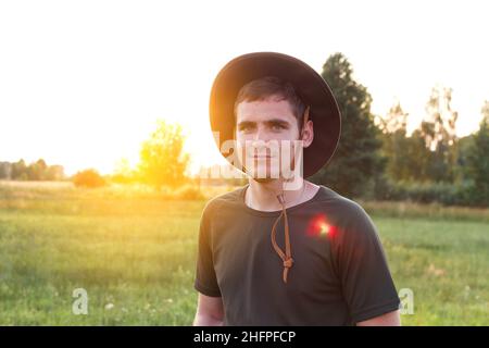 Junger Mann Bauer in Cowboy Hut auf landwirtschaftlichem Feld bei Sonnenuntergang mit Sonneneinstrahlung. Porträt eines tausendjährigen Mannes mit Hut, der auf Grasgrund steht Stockfoto