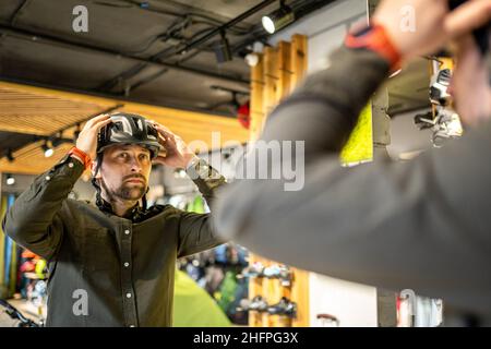 Mann, der Fahrradhelme im Sportgeschäft untersucht und sich im Spiegel ansieht. Neue Sporthelme im Fahrradladen ausprobieren. Mann setzt Fahrradhelm im Geschäft an Stockfoto