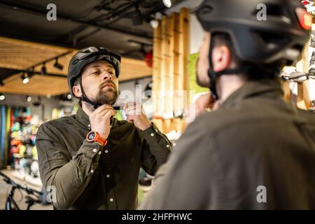 Mann, der Fahrradhelme im Sportgeschäft untersucht und sich im Spiegel ansieht. Neue Sporthelme im Fahrradladen ausprobieren. Mann setzt Fahrradhelm im Geschäft an Stockfoto