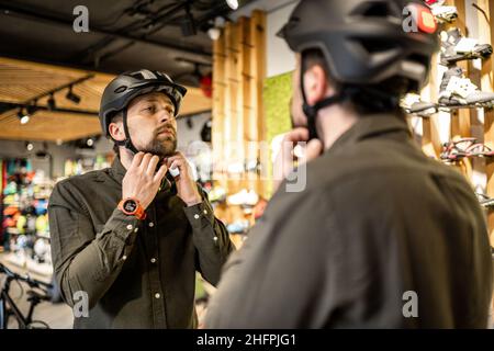 Mann, der Fahrradhelme im Sportgeschäft untersucht und sich im Spiegel ansieht. Neue Sporthelme im Fahrradladen ausprobieren. Mann setzt Fahrradhelm im Geschäft an Stockfoto