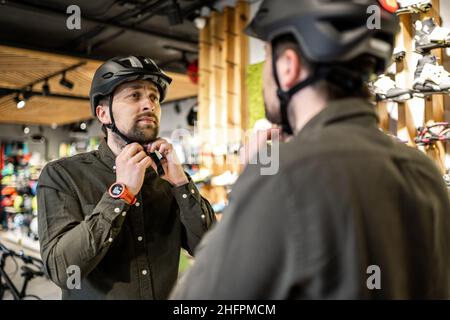 Mann, der Fahrradhelme im Sportgeschäft untersucht und sich im Spiegel ansieht. Neue Sporthelme im Fahrradladen ausprobieren. Mann setzt Fahrradhelm im Geschäft an Stockfoto