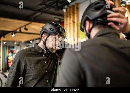 Mann, der Fahrradhelme im Sportgeschäft untersucht und sich im Spiegel ansieht. Neue Sporthelme im Fahrradladen ausprobieren. Mann setzt Fahrradhelm im Geschäft an Stockfoto