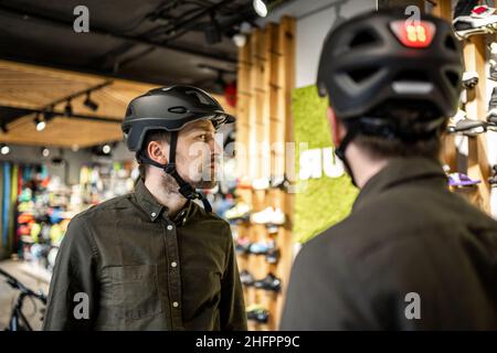 Mann, der Fahrradhelme im Sportgeschäft untersucht und sich im Spiegel ansieht. Neue Sporthelme im Fahrradladen ausprobieren. Mann setzt Fahrradhelm im Geschäft an Stockfoto