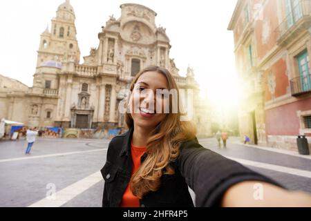 Lächelndes Mädchen in Murcia, das Selfie-Foto mit der Kathedrale Kirche Santa Maria und Stadtbild im Hintergrund gemacht hat. Mädchen porträtiert sich in Murcia Stockfoto