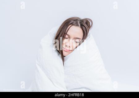 Eine hübsche Brünette, eingewickelt in eine weiße Decke, sitzt auf dem Bett. Mit einer warmen Decke warm halten. Faulheit. Stockfoto