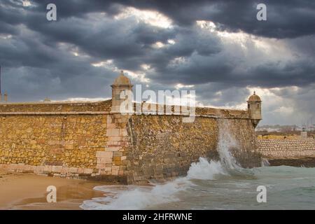 Wellen brechen an den Wänden der Festung Ponta da Bandeira, die Teil der Hafenfestungen von Lagos, Algarve, Portugal ist. Dunkle, bedrohliche Wolken. Stockfoto