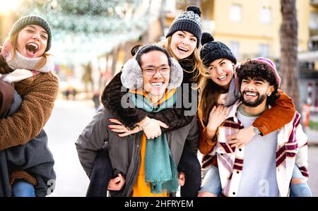 Fröhliche multikulturelle Freunde, die mit dem Huckepack am Winterreiseort spazieren gehen - Alltagsstilkonzept mit fröhlichen Jungs und Mädchen, die Spaß haben Stockfoto