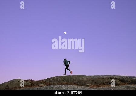 Rüden-Jogger läuft bei Dämmerung mit dem Mond entlang des felsigen Gipfels Stockfoto