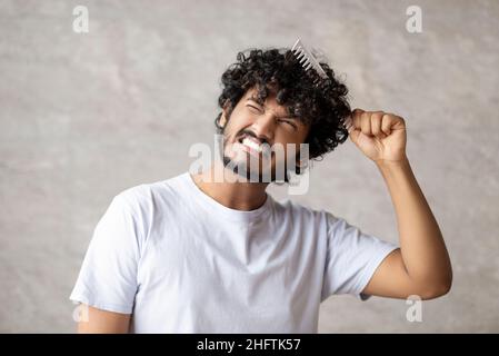 Trockenes und mattiertes Haar. Indischer junger Mann in weißem T-Shirt kombiniert mit Pinsel und Schmerzen von verworrenen lockigen Haaren Stockfoto