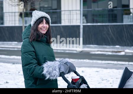 Stilvolle junge Mutter mit einer Tasse Kaffee auf einem Spaziergang mit einem Kinderwagen im Winter. Stockfoto