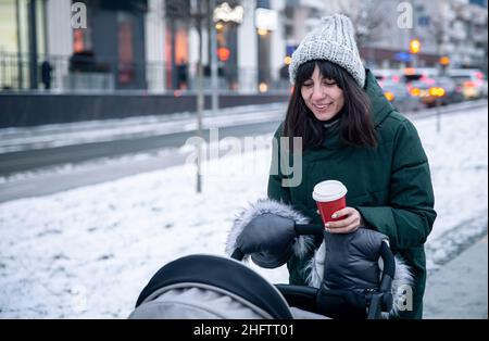 Stilvolle junge Mutter mit einer Tasse Kaffee auf einem Spaziergang mit einem Kinderwagen im Winter. Stockfoto