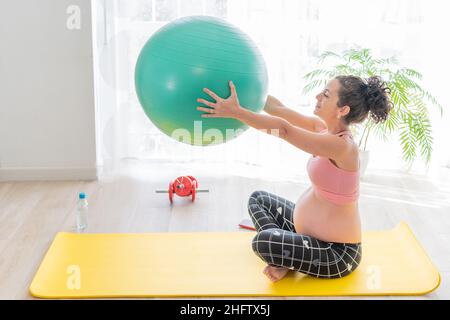 Lächelnde, hübsche junge Schwangerin, die einen grünen Fitnessball in den Armen hält und auf einer Trainingsmatte sitzt, während sie eine Flasche Wasser an ihrer Seite für Hydrati hält Stockfoto