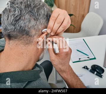 Älterer Mann, der ein Hörgerät auf sein Ohr legt, während er seinen Arzt in der Hörklinik besucht. Hörlösungen für ältere Menschen mit Gehörlosigkeit Stockfoto