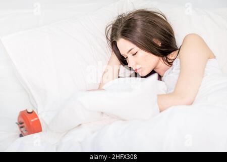 Die Brünette schläft auf einem weißen Bett. Ein süßes Mädchen im Schlafanzug liegt mit geschlossenen Augen auf dem Bett, ein Wecker liegt in der Nähe. Guten Morgen. Entspannung. Stockfoto