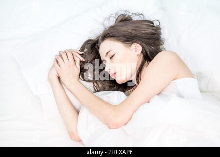 Entspannung. Guten Morgen. Die Brünette schläft auf einem weißen Bett. Ein süßes Mädchen im Schlafanzug liegt mit verschütteten Augen auf dem Bett. Schlafzimmer. Stockfoto