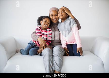 Porträt einer fürsorglichen afrikanischen Mutter, die ihre zwei niedlichen Töchter umarmt, die auf dem weißen, gemütlichen Sofa sitzen. Konzept der Familie, Liebe und Lebensstile. Stockfoto