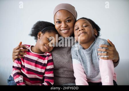 Porträt der fürsorglichen islamischen afrikanischen Mutter in Hijab umarmt ihre zwei niedlichen Töchter, die zusammen auf dem weißen gemütlichen Sofa sitzen. Konzept der Familie, Liebe und Lebensstile. Stockfoto