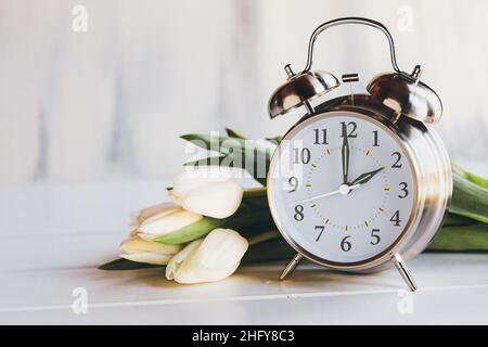 Konzept der Sommerzeit. Stellen Sie Ihre Uhren und auf 2 Uhr mit diesem Bild eines Weckers mit weißen Tulpenblüten ein. Selektiver Fokus Stockfoto