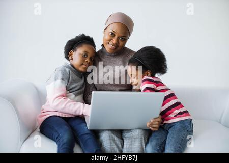Fürsorgliche afroamerikanische muslimische Mutter, die mit ihren zwei niedlichen Töchtern auf der weißen Couch sitzt und einen tragbaren Laptop benutzt. Familienzeit mit moderner Technik. Stockfoto